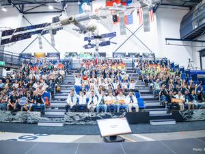 Teilnehmerinnen und Teilnehmer des internationalen CanSat-Finales jubeln auf der Tribüne im ESA-Forschungszentrum ESTEC unter Modellen von Satelliten und Raumstation