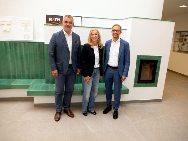 Drei Personen stehen vor einem Kachelofen und einer grünen Sitzbank.
